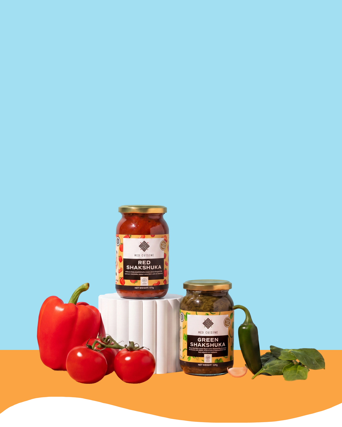 Two jars of Samadhi's Red and Green Chilli Shishmkra are displayed on a white pedestal with fresh ingredients (bell pepper, tomatoes, garlic, chili, mint) on an orange and light blue background.